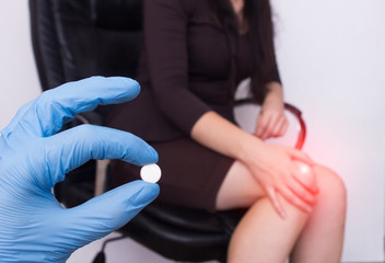 Business girl in the office sits in a computer chair and holds her knee, pain and inflammation in the knee, the doctor holds the pill in her hand