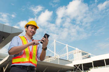 Young engineer wearing yellow safety helmet using a mobile phone in front construction building.