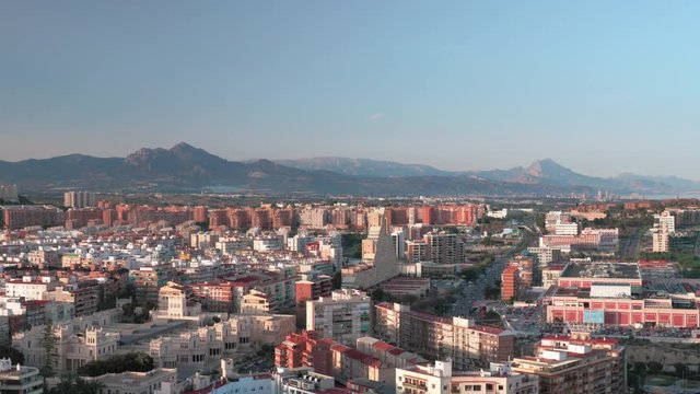 An aeral view of Alicante on a beautiful sunny day. Numerous buildings of different styles, colors and number of storeys. A wide busy street can be seen between them. There are picturesque mountains