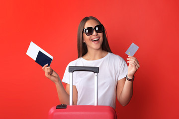 Tourist girl in summer showing plastic credit card, with sunglasses, red suitcase, passport isolated on red background.