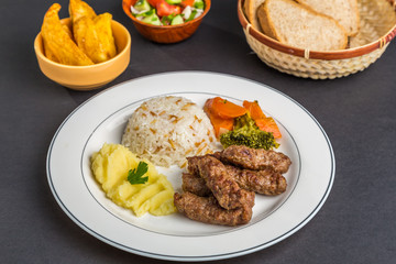 Turkish Kofte on a plate with rice, potatoes and vegetables, accompanied by bread, French fries and salad, on a black background