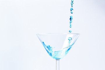 Cocktail with blue liquid in glass. Glass with blue water pouring with liquid with splashes and drops. Martini glass filling with alcohol with splashes on white background. Refreshing drink concept.
