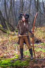 Outdoor portrait of young beautiful demon woman with ram horns, bow and arrows in forest.