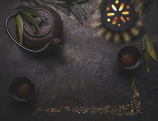 Dark tea background with black iron asian teapot and mug of hot tea on rustic table, top view,...