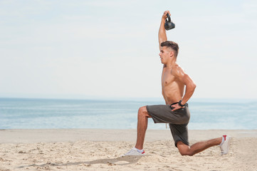 Side view of young strong man training with kettle bell. Muscular male athlete exercising outdoors at the beach against sky. Cross strength training, place for text
