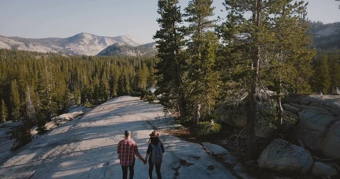 Aerial tracking shot of young newlyweds hiking together on amazing white rock towards epic Yosemite park forest skyline.