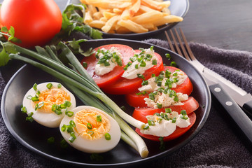 Half boiled eggs with tomatoes, green onions, sprinkled with greens in a black plate on a gray wooden table. Close-up