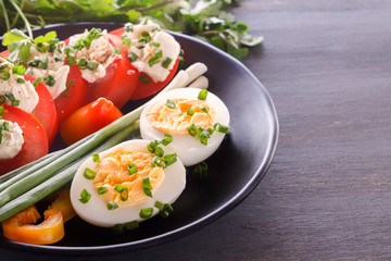Half boiled eggs with tomatoes, green onions, sprinkled with greens in a black plate on a gray wooden table. Close-up. Copy space