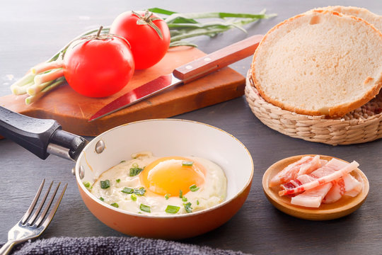 Fried egg in a pan, sliced bread, bacon, tomatoes and green onions cooked for breakfast on a wooden gray table.