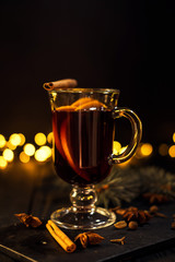 cinnamon lies in a glass, closeup glass of mulled wine with orange and cinnamon on dark black background,  Christmas tree and lights, large yellow bokeh, mulled wine set
