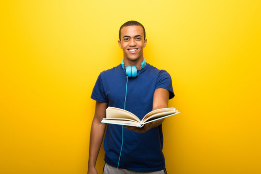 African american man with blue t-shirt on yellow background holding a book and giving it to someone