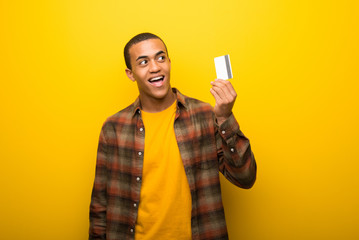 Young african american man on vibrant yellow background holding a credit card and thinking