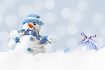 Christmas happy snowman wearing blue hat and scarf with xmas balls on abstract lights bokeh background, white snowflakes falling on winter landscape, merry Christmas and -happy new year greeting card