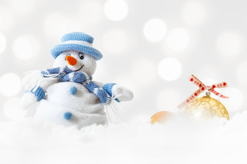 Happy funny snowman wearing blue hat and scarf with xmas balls on abstract lights background, white soft snowflakes fall on winter landscape, merry Christmas and happy new year greeting card concept