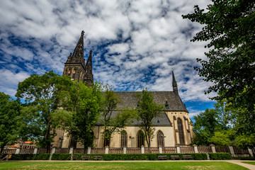 Amazing overcast summer day landscape view of the St. Peter and St. Paul Basilica, Prague, Czech Republic, colorful side view with trees, bushes and empty brown wooden benches in green public park