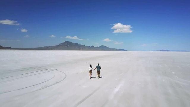 Drone follows romantic couple holding hands, running towards epic white flat land of Utah salt desert lake, having fun.