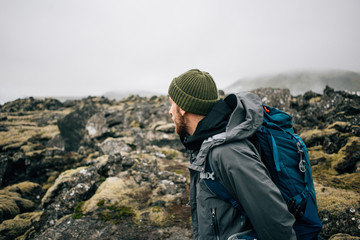 Young man in hiking trekking gear, waterproof jacket, green knit beanie and hike backpack walk through moss covered rough iceland terrain. Explore travel real wilderness lifestyle