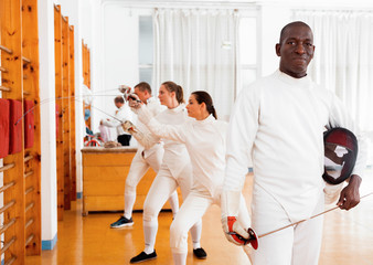 Active african american male fencer in uniform standing with mask and foil at fencing room