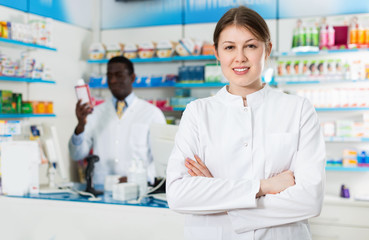 Experienced female pharmacist standing on background with male colleague in pharmacy