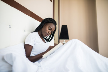 Young african american woman relaxing at home and reading a book lying on the bed