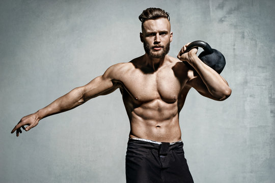 Young muscular man training with kettlebells. Photo of man with naked torso on grey background. Strength and motivation