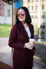 Brunette young caucasian woman in casual jacket or coat and sunglasses walking european city streets, smiling happily with a cup of coffee. Lifestyle portrait indoors.