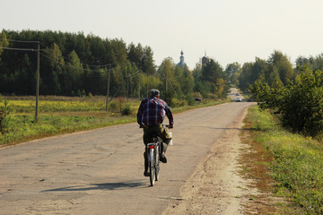 Obraz premium Old man drives bicycle in the village road with the background of fall forest, church in sunny day