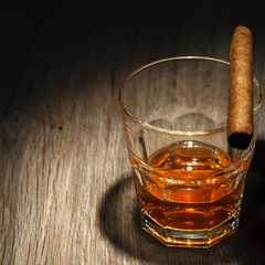 A glass of alcoholic beverage with ice cubes and a cigar on a wooden table. Whiskey in glass