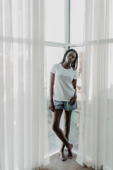Morning of beautiful African-American woman standing near window in curtains