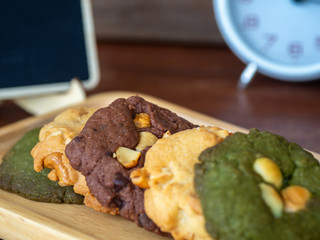 Multiple color cookies inclusive peanut butter, green tea cookies, and Chocolate Chip Cookies. Overlaid by alternating colors on wood plate. White clock and blackboard placed in the back side
