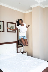 Fototapeta na wymiar Full length portrait of a cheerful afro american woman in earphones singing and jumping on bed indoors