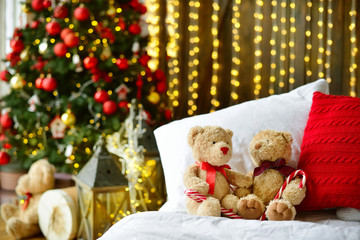 Two teddy bears siting on the bed with red candys near christmas tree.