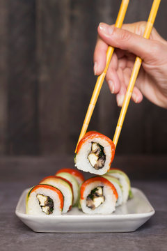 Hand picking up sushi rolls from white plate by bamboo chopsticks,  on gray table, dark wooden background. Culinary, menu, asian food concept. Close-up, copy space, layout design, vertical format