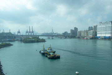 The barge is floating on the sea. Long bridge across the water. Construction cranes. Tall city buildings, waterfront. City by the sea Hong Kong.