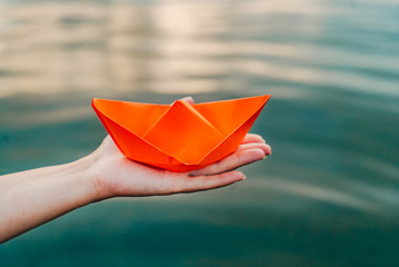 An orange paper ship in a female hand over the water. Woman's hand holding paper boat above the water. Close-up