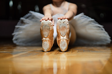 Ballerina sitting on the floor of classroom and and holding for tiptoes of white ballet shoes on...