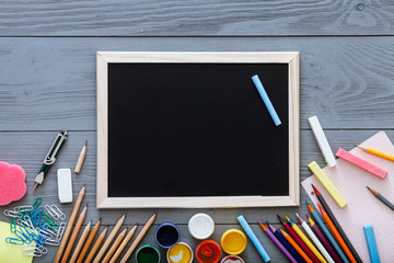 Chalkboard on grey dark desk with colorful pencils, paints, other school supplies for schoolwork, back to school sale concept, creative workplace for new learning year, top view, copy space