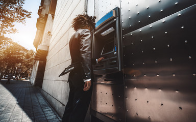 Wide-angle view of a curly businessman in a formal suit using an outdoor cash dispenser to increase...