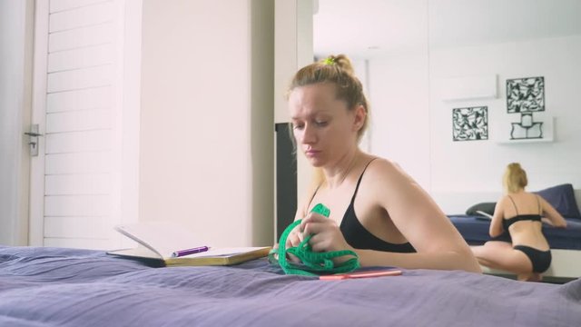 The concept of overweight and weight loss. A woman measures herself with a measuring tape in the bedroom, Looking at herself in the mirror, and writes the results into a notebook