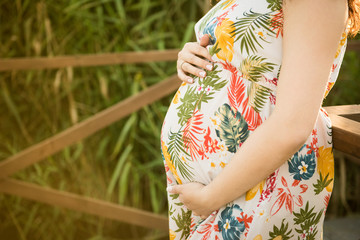 A cropped look at a young woman standing in a sunlit forest; she is very pregnant and cradles her rounded belly gently