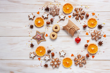 Christmas holiday  wreath  background with gift box, homemade cookies and gingerbread,  dry orange slices, nuts, spices and cones on white wooden background. Top view, close up