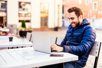 Smiling young man working from outdoor office. Man wearing jacket typing on laptop on outdoor terrace in urban environment