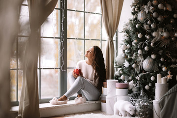 Charming dark-haired girl dressed in pants, sweater and warm slippers holds a red cup sitting on...