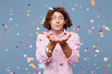 Portrait of man happy standing around falling down confetti, over blue background. Young man blowing confetti off his hands.