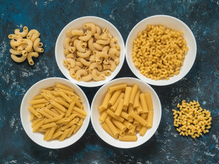 Various dry pasta in white plates on a blue grunge background. Top view. Copy space