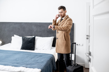 Dressed businessman standing with a suitcase and calling phone in the hotel room before the check...