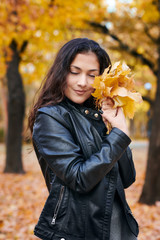 Pretty woman is posing with bunch of maple's leaves in autumn park. She's closing eyes and dreaming. Beautiful landscape at fall season.