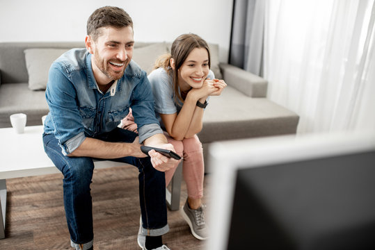 Young lovely couple with happy emotions watching TV sitting together on the table at home - Powered by Adobe
