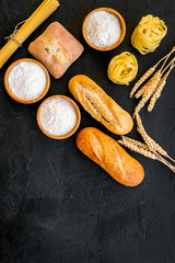 Farinaceous food. Fresh bread and raw pasta near flour in bowl and wheat ears on black background top view space for text