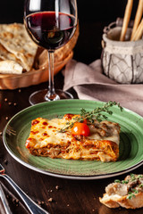 The concept of Italian cuisine. Baked lasagna with minced bolognese, pasta, cherry tomatoes lies on a green plate in a restaurant. A glass of red wine and bread basket on the table